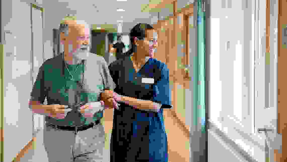 Smiling senior man walking arm in arm with female nurse while looking away in alley at nursing home