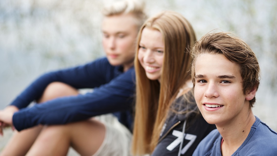 Tre tonåringar sitter på rad och skrattar. En pojke tittar in i kameran och ler.