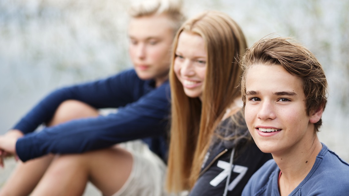 Tre tonåringar sitter på rad och skrattar. En pojke tittar in i kameran och ler.