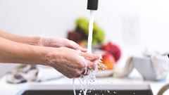 En person tvättar händerna under rinnande vatten från vattenkran i kök.

