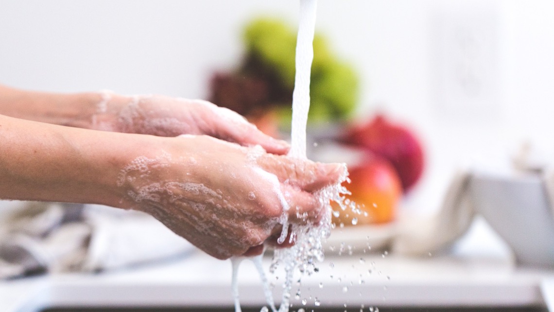 En person tvättar händerna under rinnande vatten från vattenkran i kök.

