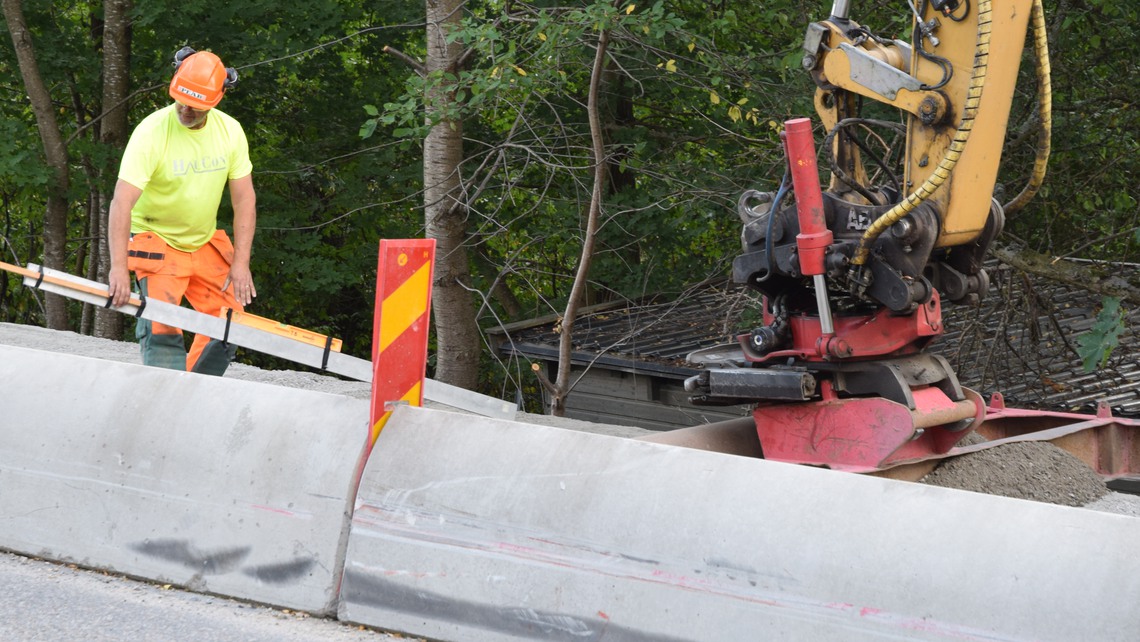 Vägarbetare arbetar bredvid en väg intill en grävskopa.