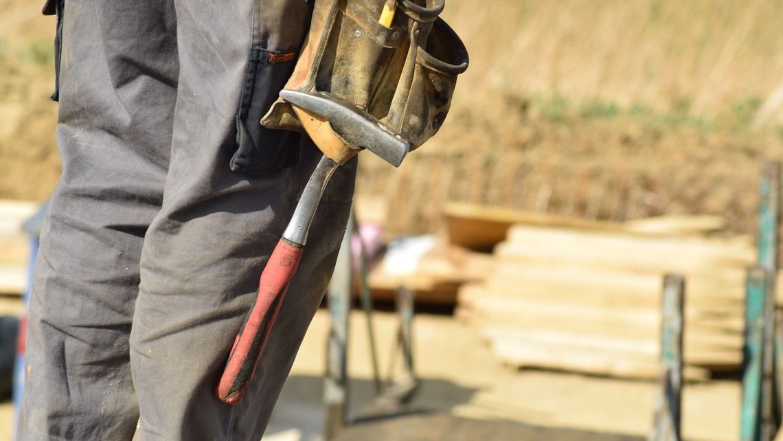 En man i arbetskläder står vid en arbetsplats och har en hammare hängandes på byxan. 