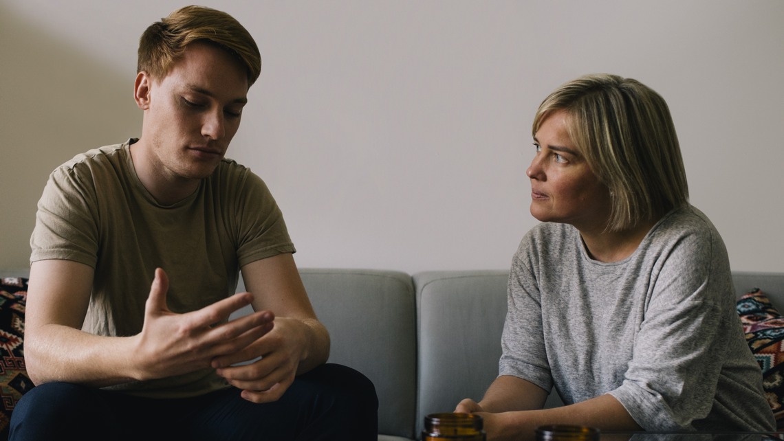 Mature parent looking at sad son sitting on sofa at home