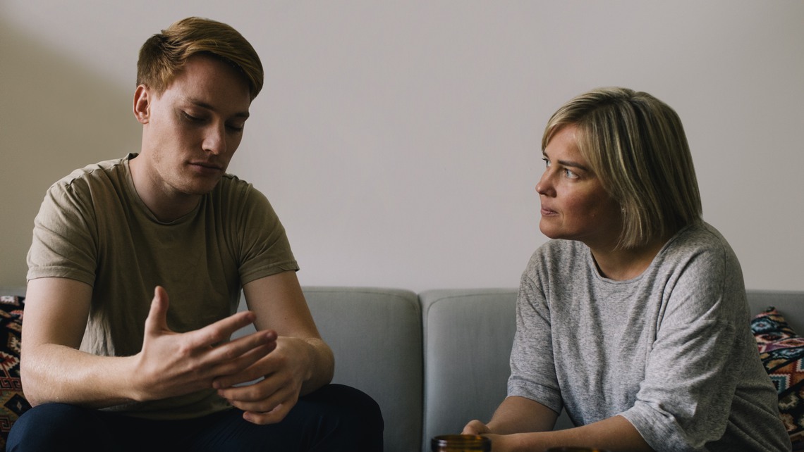 Mature parent looking at sad son sitting on sofa at home