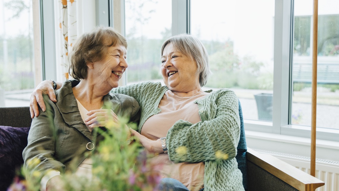 Smiling elderly women sitting with arm around while looking at each other on sofa against window at retirement home