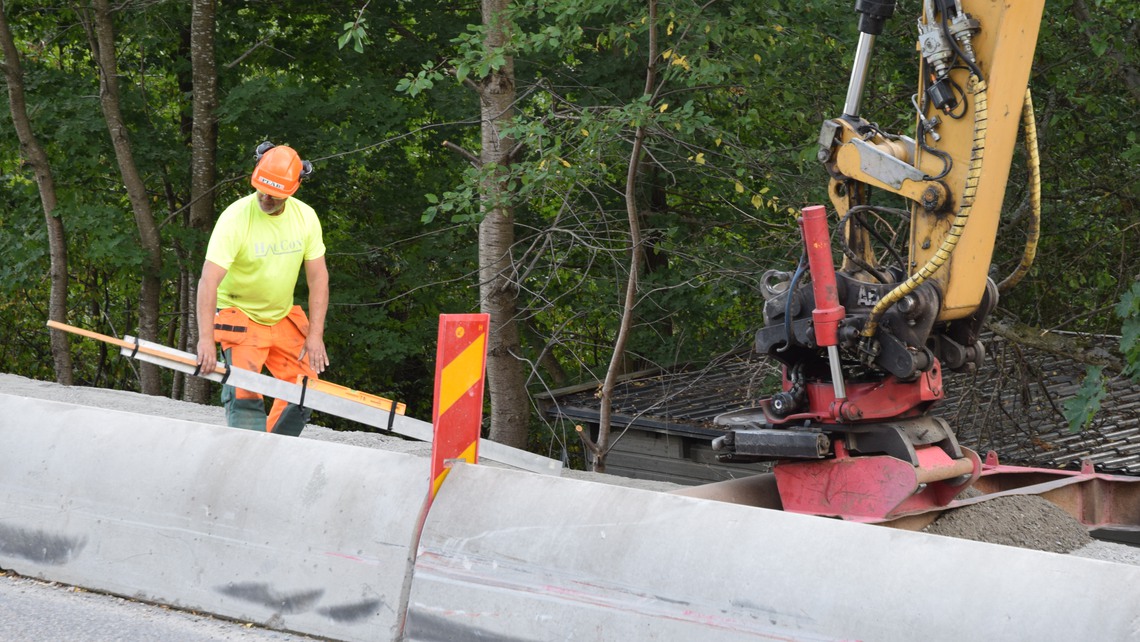 Vägarbetare arbetar bredvid en väg intill en grävskopa.