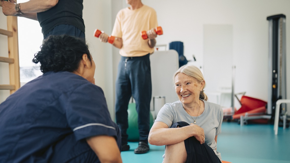 Smiling senior woman stretching and sitting on exercise mat while talking with therapist in rehabilitation center