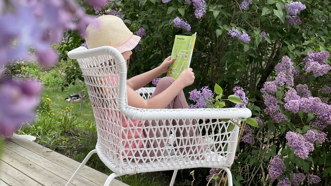 Flicka sitter med solhatt och läser omgiven av syréner.