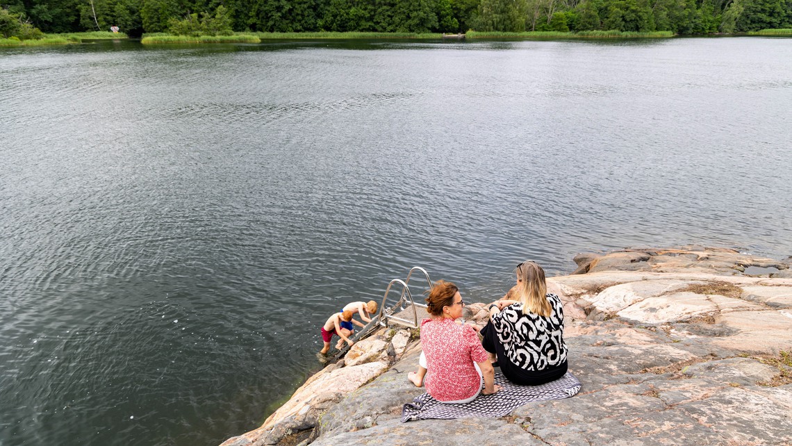 Två barn kliver upp från badstege och två kvinnor sitter på klippor ovanför.