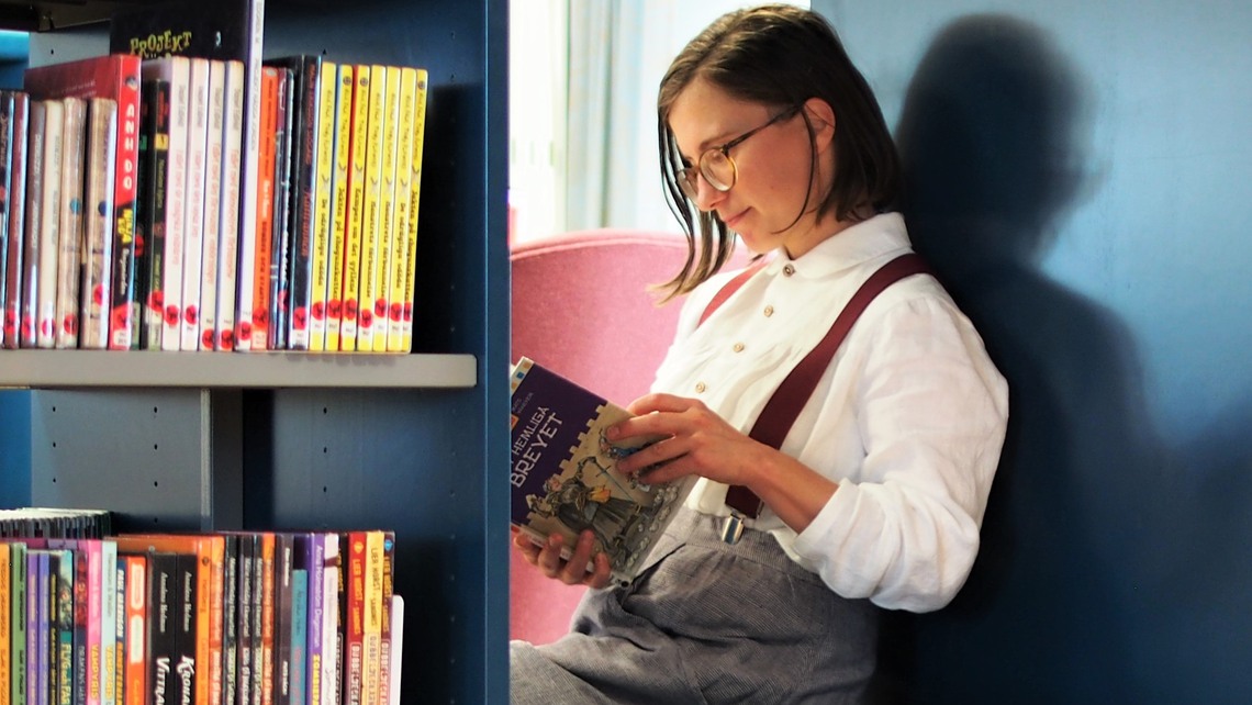 Kvinna med glasögon och hängslen sitter i en bibliotekshörna med blå hylla och läser.