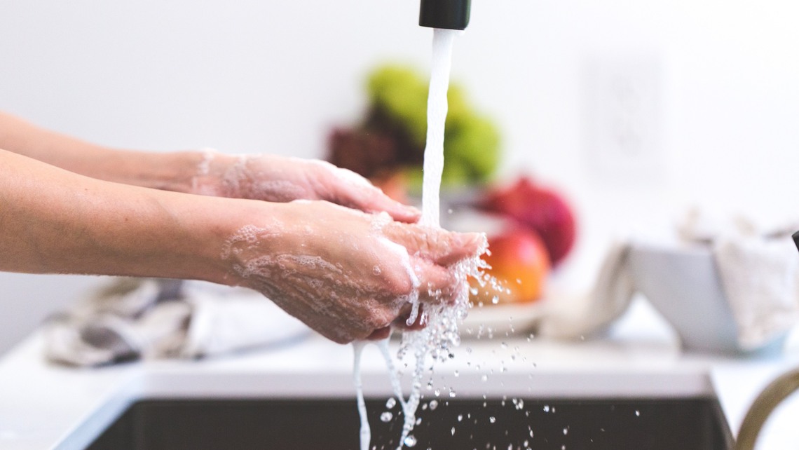 En person tvättar händerna under rinnande vatten från vattenkran i kök.