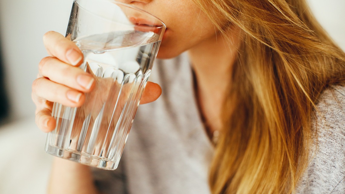 En kvinna med blont hår och grå t-shirt dricker ur ett glas med vatten. Man ser bara halva hennes ansikte.