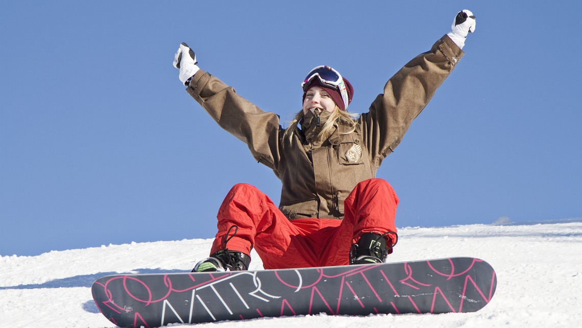 Ung tjejer med en snowboard sitter i snön med armarna uppsträckta.